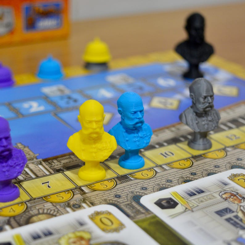 May include: A board game with colorful miniature busts of historical figures. The game board features a blue and gold design with numbered spaces. The busts are in yellow, blue, purple, black, and gray. Cards with illustrations are at the bottom.
