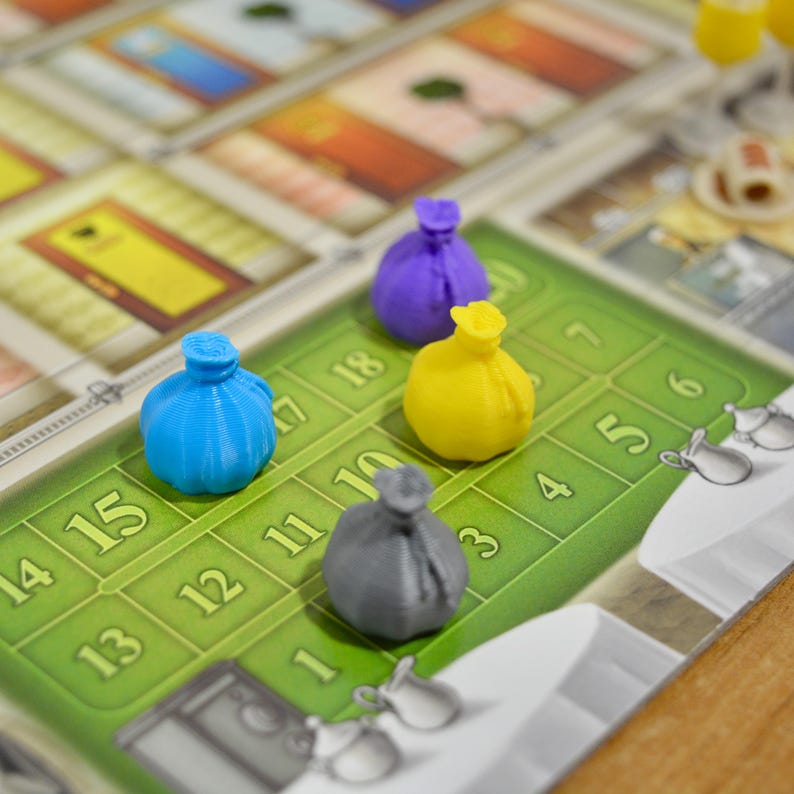 May include: Four small, three-dimensional plastic game tokens shaped like tied sacks in blue, purple, yellow, and gray. These tokens are positioned on a green board game track featuring numbered squares from one to eighteen.