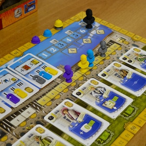 May include: A board game featuring a blue and yellow game board with numbered tracks, small colored plastic busts, and rectangular playing cards depicting historical figures and numerical values. The board includes sections labeled A, B, and C.