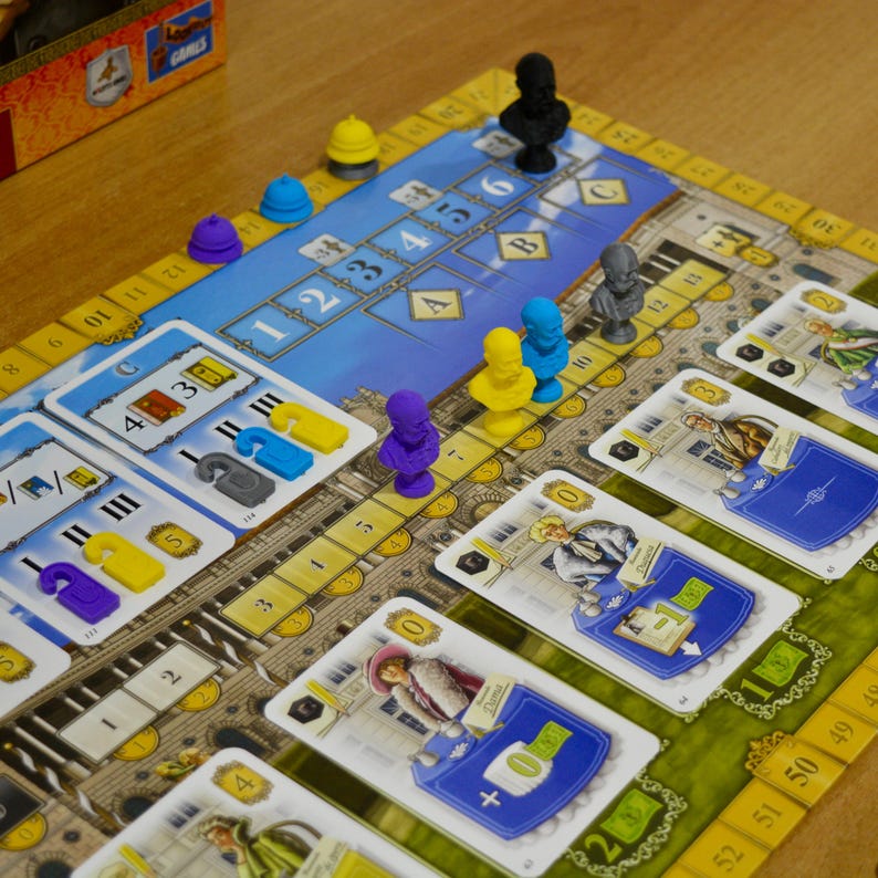 May include: A board game featuring a blue and yellow game board with numbered tracks, small colored plastic busts, and rectangular playing cards depicting historical figures and numerical values. The board includes sections labeled A, B, and C.