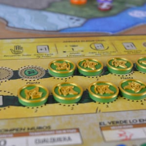 May include: A board game in a close-up shot. The game board is yellow with green and brown accents. There are several green and gold tokens with a plant design. The board has text in Spanish, including "Planta" and "El Verde Lo Envuelve Todo."