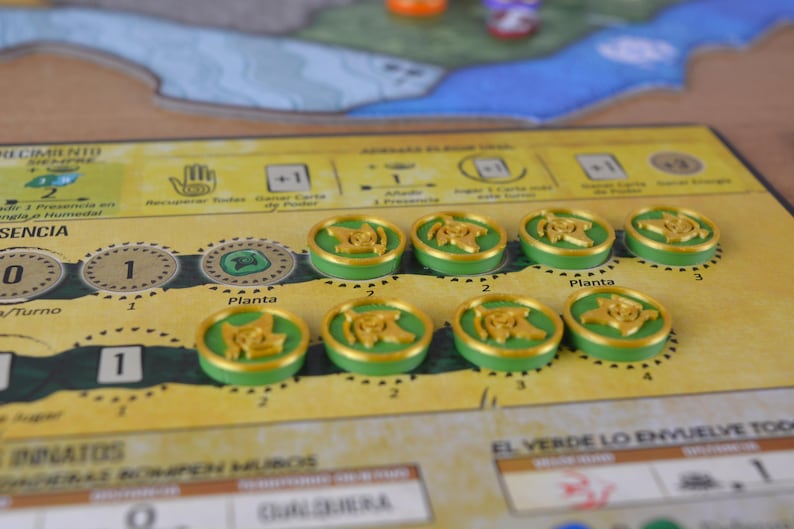May include: A board game in a close-up shot. The game board is yellow with green and brown accents. There are several green and gold tokens with a plant design. The board has text in Spanish, including "Planta" and "El Verde Lo Envuelve Todo."