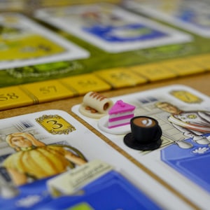 May include: Close-up of a board game in progress. The game features cards with illustrations of people and numbers. Miniature food items, including a cake slice and a coffee cup, are placed on the cards, indicating game pieces. The game board has a yellow and green color scheme.