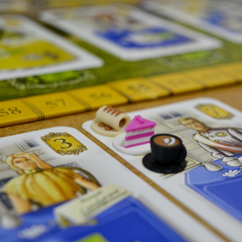 May include: Close-up of a board game in progress. The game features cards with illustrations of people and numbers. Miniature food items, including a cake slice and a coffee cup, are placed on the cards, indicating game pieces. The game board has a yellow and green color scheme.