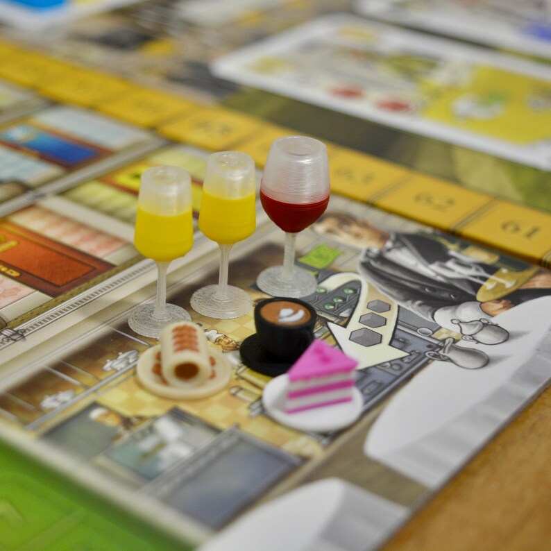 May include: A board game scene featuring miniature food and drink items. There are two yellow drinks, a red wine, a coffee, a cake slice, and a pastry on a plate. The game board has a yellow and brown color scheme.