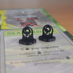 May include: Two black, 3D-printed game pieces shaped like stylized tombstones. The pieces have a circular design at the top and are set on a game board with text in Spanish. The background is blurred.