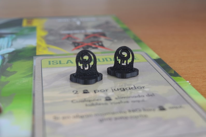 May include: Two black, 3D-printed game pieces shaped like stylized tombstones. The pieces have a circular design at the top and are set on a game board with text in Spanish. The background is blurred.