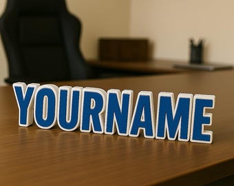 May include: A three-dimensional desk sign featuring the word YOURNAME in bold, capital letters. The letters have a deep blue front face with a thick white border, standing upright on a flat surface to display the custom text clearly.