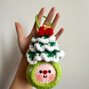 May include: A handmade Christmas ornament featuring a green and white crocheted tree design atop a round, green face with rosy cheeks, a red nose, and a small mouth. The ornament has a green loop for hanging and is adorned with a red bow and white pearls.