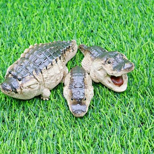 May include: Three detailed alligator figurines with textured skin and open mouths rest on a bed of green artificial grass. The alligators are in various sizes and poses, showcasing their realistic features.