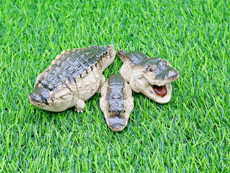 May include: Three detailed alligator figurines with textured skin and open mouths rest on a bed of green artificial grass. The alligators are in various sizes and poses, showcasing their realistic features.
