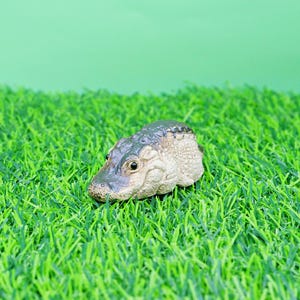 May include: A miniature crocodile head figurine with a textured, pale body and a dark green head. It is placed on a bed of bright green artificial grass, set against a light green background.