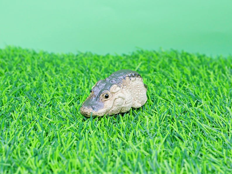 May include: A miniature crocodile head figurine with a textured, pale body and a dark green head. It is placed on a bed of bright green artificial grass, set against a light green background.