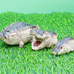 May include: Three detailed alligator figurines of different sizes are arranged on artificial green grass. The largest alligator is closed-mouthed, the middle one has its mouth open, revealing a red interior, and the smallest is also closed-mouthed.