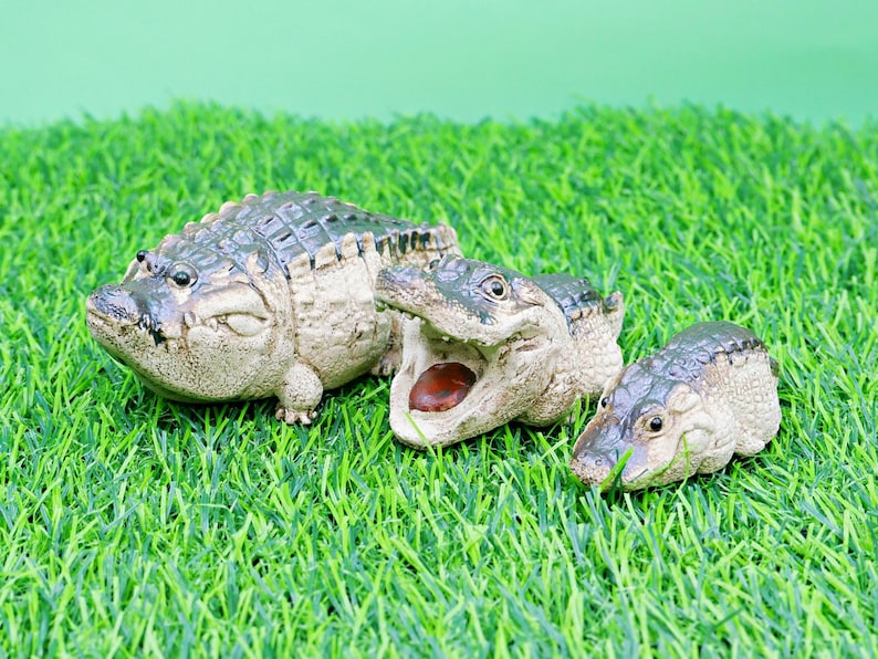 May include: Three detailed alligator figurines of different sizes are arranged on artificial green grass. The largest alligator is closed-mouthed, the middle one has its mouth open, revealing a red interior, and the smallest is also closed-mouthed.