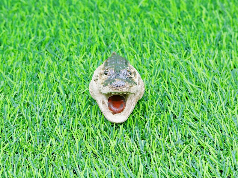 May include: A detailed alligator head figurine, with a green and tan colour scheme, set against a backdrop of vibrant green artificial grass. The alligator's mouth is open, revealing a red interior.