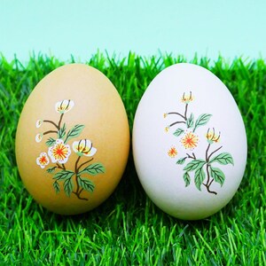 May include: Two decorative eggs with floral designs. One egg is tan, the other is white. Each egg features a branch with white and orange flowers and green leaves. The eggs are on a bed of green grass.