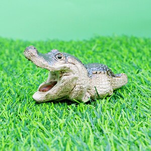 May include: A detailed figurine of an alligator with its mouth open, showcasing its teeth and tongue. The alligator is primarily beige and green, set against a backdrop of green artificial grass and a light green background.