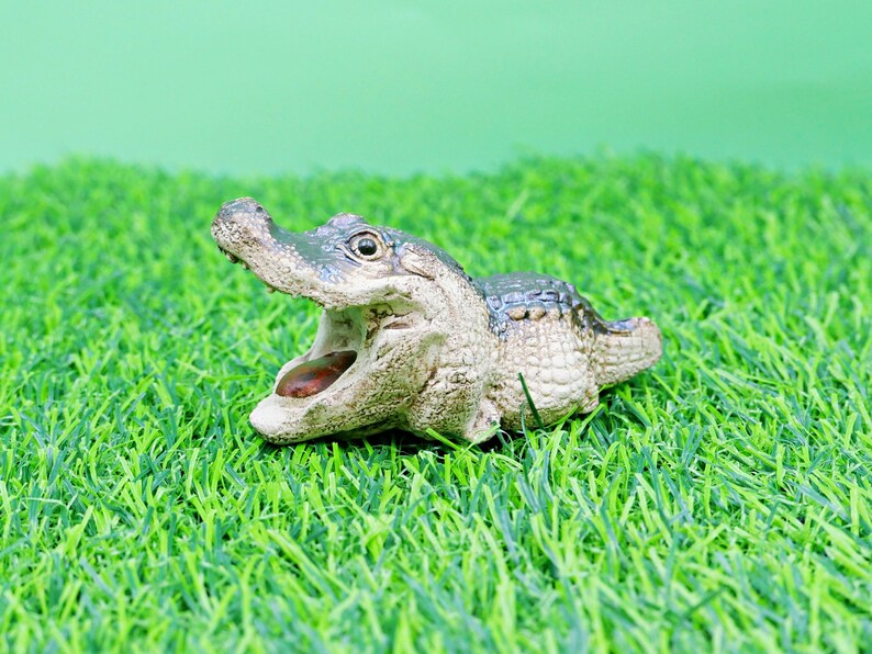 May include: A detailed figurine of an alligator with its mouth open, showcasing its teeth and tongue. The alligator is primarily beige and green, set against a backdrop of green artificial grass and a light green background.