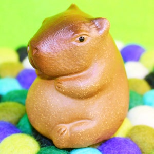 May include: A brown ceramic figurine of a capybara sitting on a colourful background of felt balls.