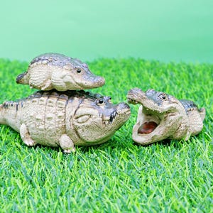 May include: Three ceramic alligator figurines on artificial green grass. One small alligator is stacked on top of a larger one. The third alligator has its mouth open, revealing a brown interior.