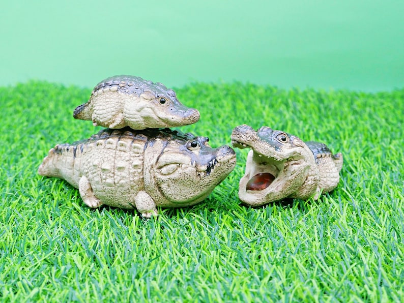 May include: Three ceramic alligator figurines on artificial green grass. One small alligator is stacked on top of a larger one. The third alligator has its mouth open, revealing a brown interior.