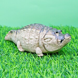 May include: A detailed figurine of an alligator, with a textured, tan body and dark grey scales. The alligator is resting on a bed of green artificial grass, with a light green background.