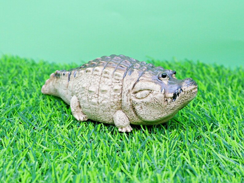 May include: A detailed figurine of an alligator, with a textured, tan body and dark grey scales. The alligator is resting on a bed of green artificial grass, with a light green background.