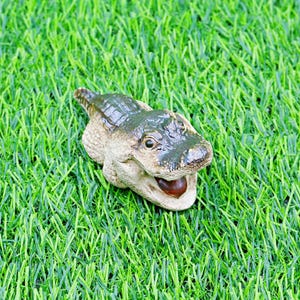 May include: A ceramic alligator figurine in green and beige tones. The alligator is positioned on a bed of green artificial grass. The alligator's mouth is open, showing a dark brown interior.