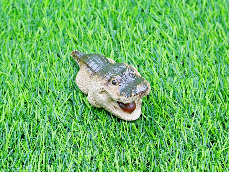 May include: A ceramic alligator figurine in green and beige tones. The alligator is positioned on a bed of green artificial grass. The alligator's mouth is open, showing a dark brown interior.