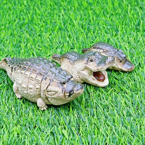 May include: Three detailed alligator figurines with textured skin and open mouths rest on a bed of green artificial grass. The alligators are in varying poses, showcasing their realistic features and colour variations.