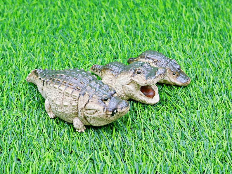 May include: Three detailed alligator figurines with textured skin and open mouths rest on a bed of green artificial grass. The alligators are in varying poses, showcasing their realistic features and colour variations.