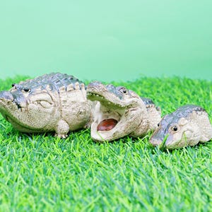 May include: Three small alligator figurines are displayed on artificial green grass against a light green background. One alligator has its mouth open. The figurines are detailed and realistic.
