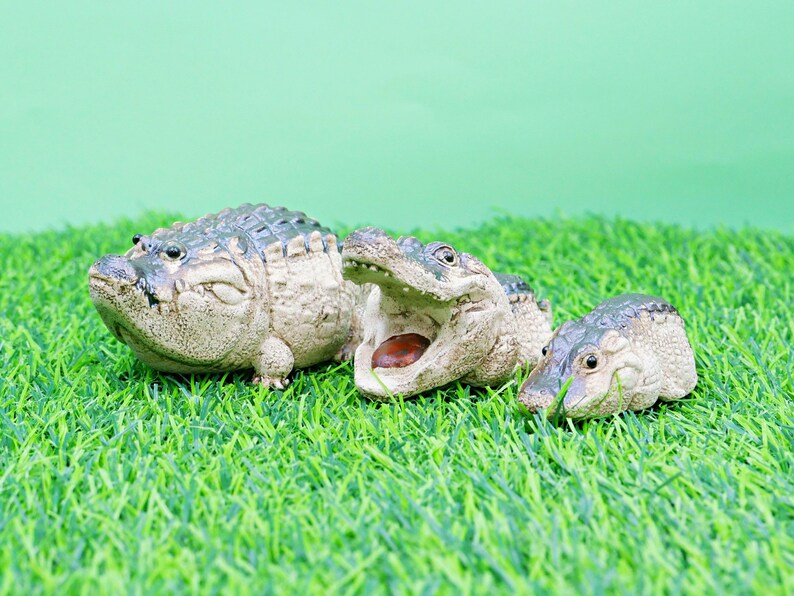 May include: Three small alligator figurines are displayed on artificial green grass against a light green background. One alligator has its mouth open. The figurines are detailed and realistic.