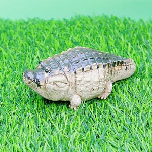 May include: A detailed figurine of an alligator, with a textured, dark green and tan body. The reptile is positioned on artificial green grass, with a light green background.