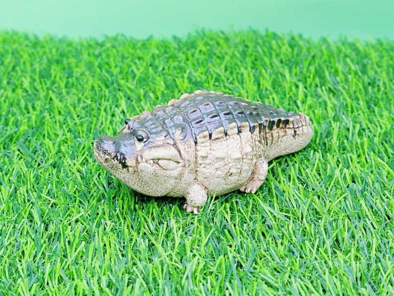 May include: A detailed figurine of an alligator, with a textured, dark green and tan body. The reptile is positioned on artificial green grass, with a light green background.