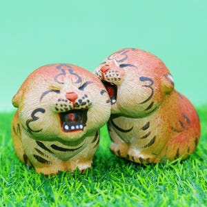 May include: Two small, hand-painted tiger figurines with orange and tan fur, black stripes, and wide-open mouths showing white teeth. The tigers are positioned side-by-side, with one slightly behind the other, both featuring rounded, expressive faces.