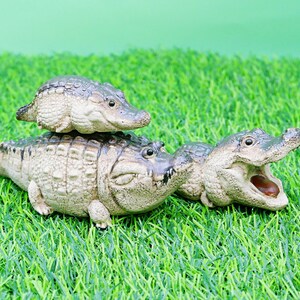 May include: Three detailed alligator figurines of varying sizes are displayed on artificial green grass. The largest alligator has its mouth open, revealing its teeth and interior. The smallest alligator is perched on top of the largest.