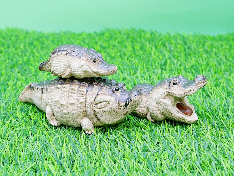 May include: Three detailed alligator figurines of varying sizes are displayed on artificial green grass. The largest alligator has its mouth open, revealing its teeth and interior. The smallest alligator is perched on top of the largest.
