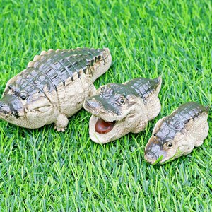 May include: Three ceramic alligator figurines of different sizes on green artificial grass. The alligators are painted in green, brown, and beige, with open mouths and detailed scales. The largest is approx. 15 cm long.