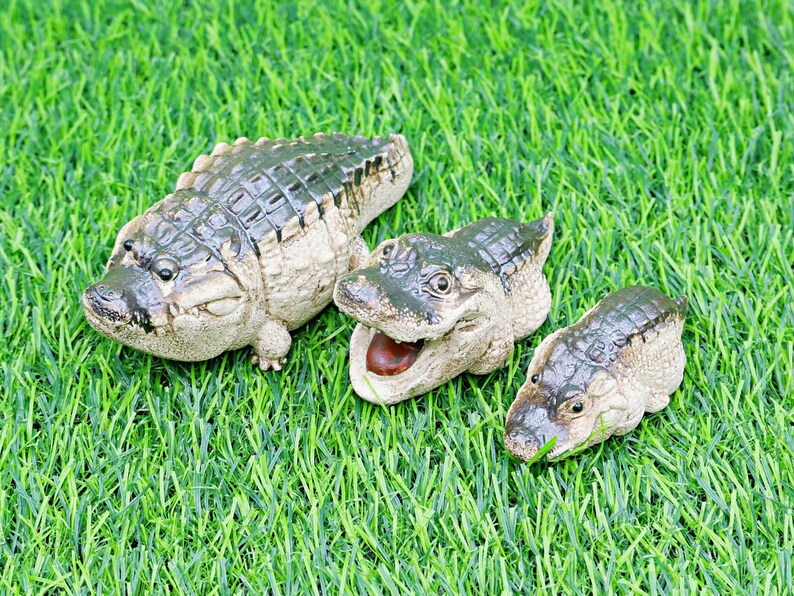 May include: Three ceramic alligator figurines of different sizes on green artificial grass. The alligators are painted in green, brown, and beige, with open mouths and detailed scales. The largest is approx. 15 cm long.