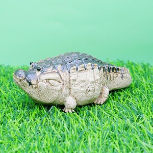 May include: A detailed figurine of an alligator, with a textured, grey-green body and a lighter beige underbelly. The alligator is resting on a bed of artificial green grass, with a light green background.