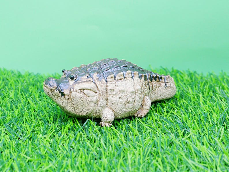 May include: A detailed figurine of an alligator, with a textured, grey-green body and a lighter beige underbelly. The alligator is resting on a bed of artificial green grass, with a light green background.