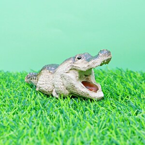 May include: A detailed figurine of an alligator with its mouth open, revealing a brown interior. The alligator is light grey and green, set against a green background and artificial grass.