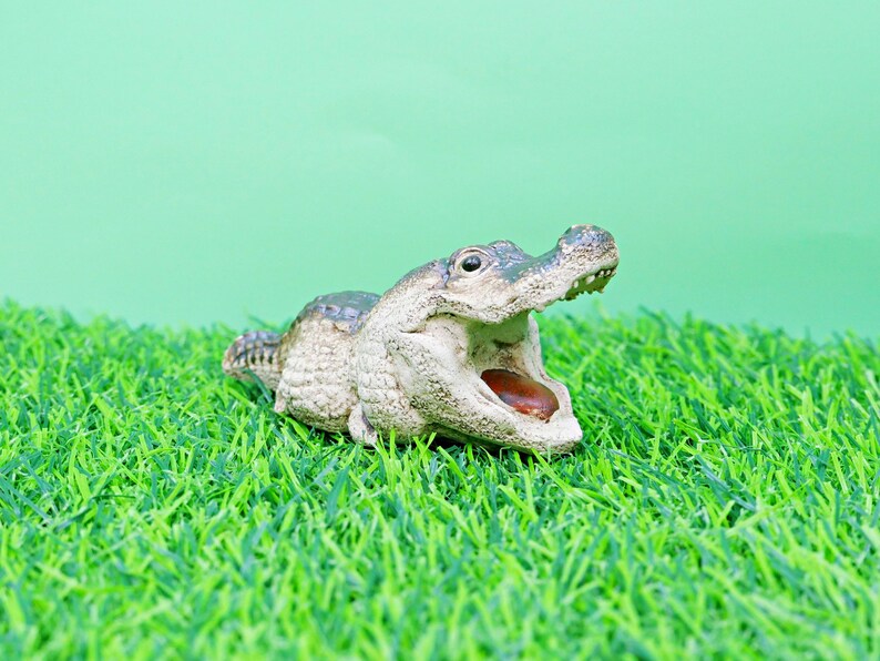 May include: A detailed figurine of an alligator with its mouth open, revealing a brown interior. The alligator is light grey and green, set against a green background and artificial grass.