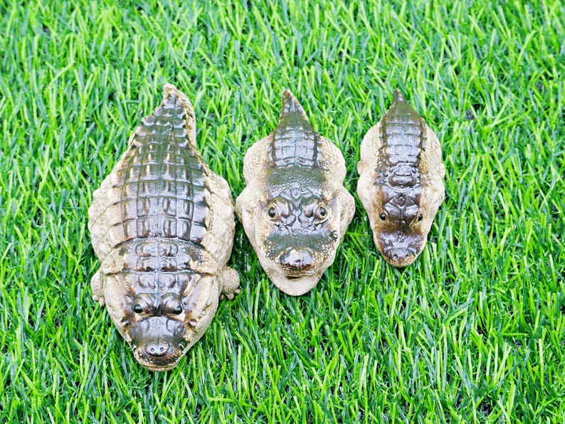 May include: Three decorative alligator figurines of different sizes, set on green artificial grass. The alligators are brown and tan, with detailed textures and realistic features, resembling miniature sculptures.
