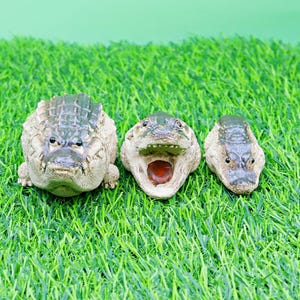 May include: Three ceramic alligator figurines with textured skin and open mouths, arranged on a bed of green artificial grass. The alligators are in varying sizes and have a neutral colour palette.
