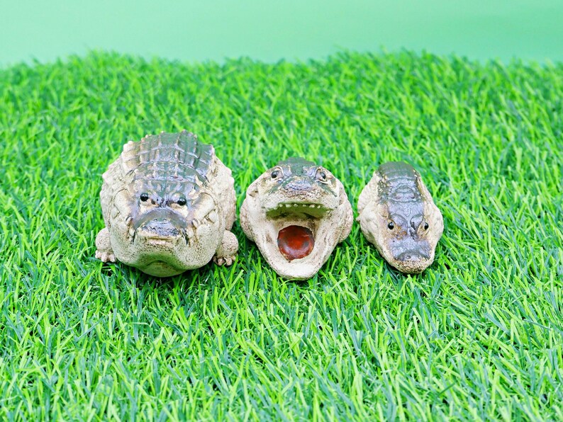 May include: Three ceramic alligator figurines with textured skin and open mouths, arranged on a bed of green artificial grass. The alligators are in varying sizes and have a neutral colour palette.