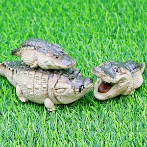 May include: Three ceramic alligator figurines of different sizes are arranged on green artificial grass. The alligators are beige with dark green detailing. One has its mouth open, showing a brown interior. The scene is set on a green lawn.