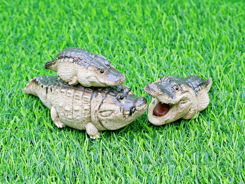 May include: Three ceramic alligator figurines of different sizes are arranged on green artificial grass. The alligators are beige with dark green detailing. One has its mouth open, showing a brown interior. The scene is set on a green lawn.